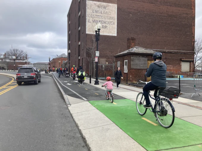 An off-street separated cycle path is shown with around 20 bikes riding in it. In the foreground is a small child (around 6-7) riding in front of an adult; further up the bike mix includes a mix of cargo bikes. There is one car pictured in the road lane to the left of the bikes.