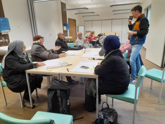 A group of a dozen or so local residents sitting around a table in a community centre. On the table are some paper maps and some felt tip pens. A young man is standing up and speaking to them while holding a coffee