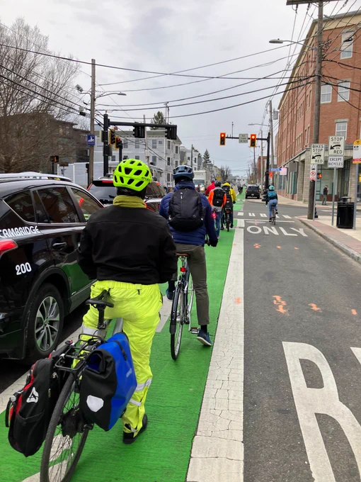 A line of around 7 bicyclists wait at a stop light in a bright green painted bike lane. There are two cars stopped at hte light to their left, and a large bus only lane to their right. They are wearing winter weather gear, and the sky is overcast.