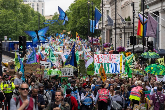 A large march of people walking down Park Lane in London. Protesters are holding banners and props including butterfly puppets