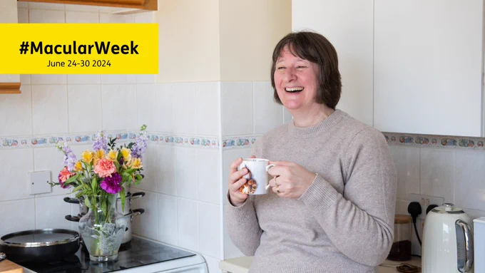 Nancy smiling as she holds a cup of tea in her kitchen. On the side is a vase of pink, purple, violet and yellow flowers. Top left is the hashtag Macular Week, with dates June 24-30 2024.