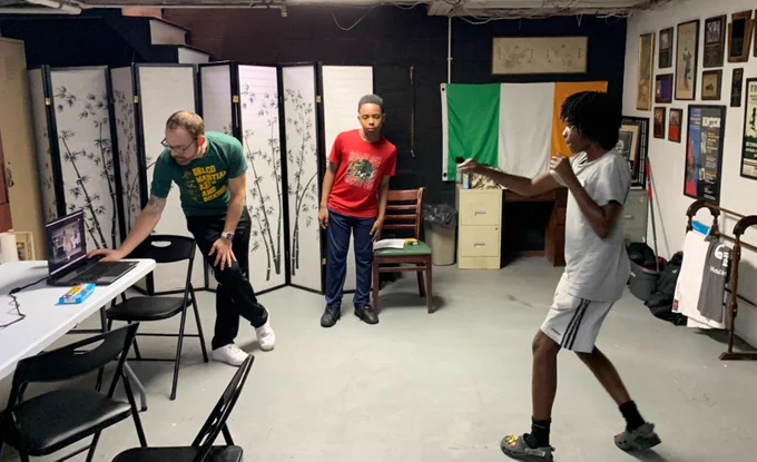 Three people in a room: one using a laptop, another watching, and a third practicing boxing. The room has a large table, chairs, a screen divider, and an Irish flag on the wall.
