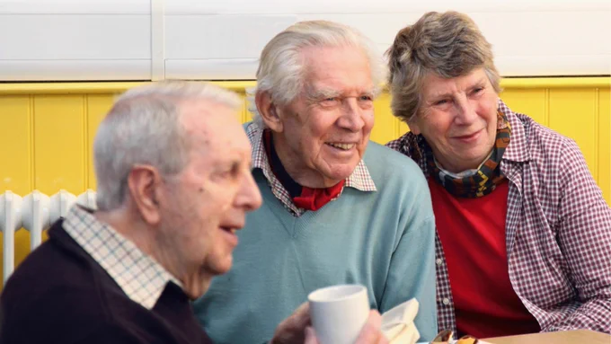 Two elderly men and one elderly woman with grey hair, sat around a table at a support group, smiling.