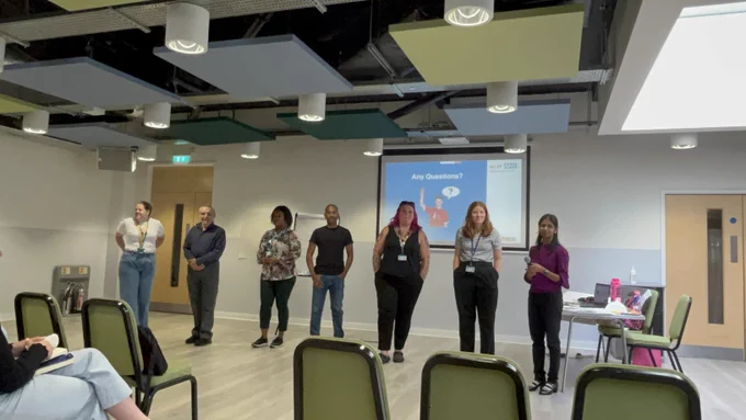 People standing at the front of a room, in front of a powerpoint display.