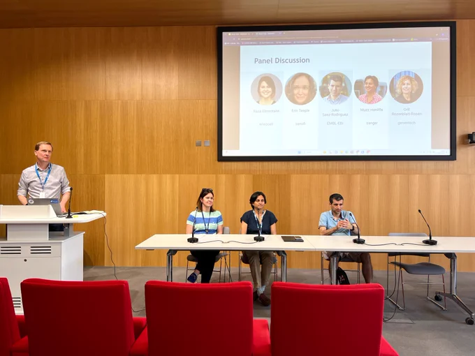 Three panellists sit at a table with microphones, listening to a question from the audience. Behind them, the screen shows the list of all panellists. To the left, David Hulcoop stands at a lectern.