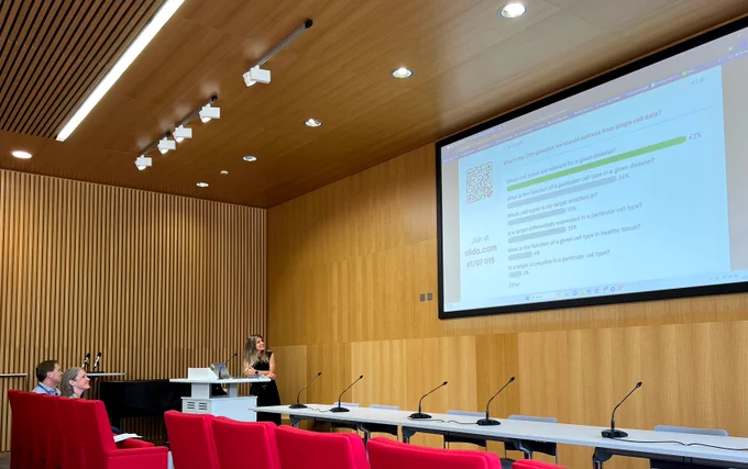 Annalisa Buniello stands at a lectern, looking back towards the screen which displays the results of an interactive poll. The photo includes the front row of the lecture theatre, with people looking up at the screen.