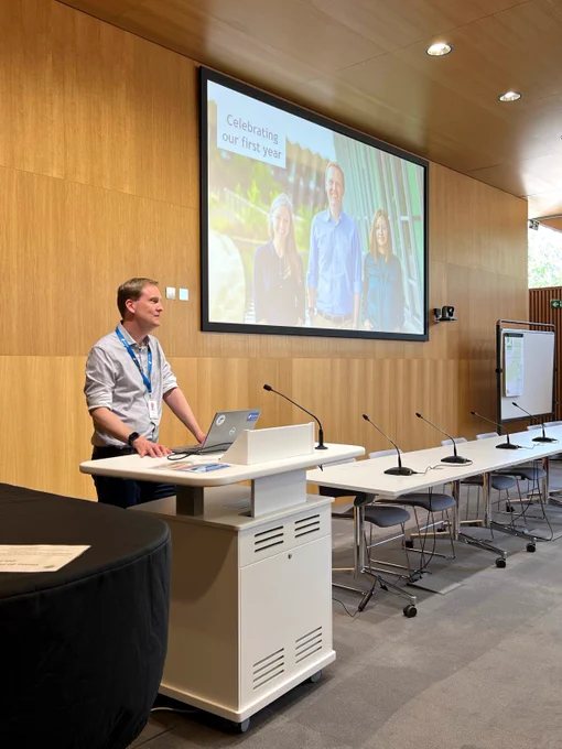 David Hulcoop stands at a lectern, facing out towards the audience and delivering a talk. The screen behind him shows a photo of the three Open Targets directors with the caption: Celebrating our first year. 