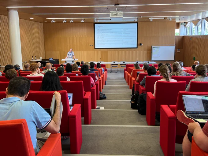 David Hulcoop stands at the front of a lecture theatre. The photo is taken from the back of the theatre so the audience can be seen looking at the projected screen which displays the agenda for the afternoon.
