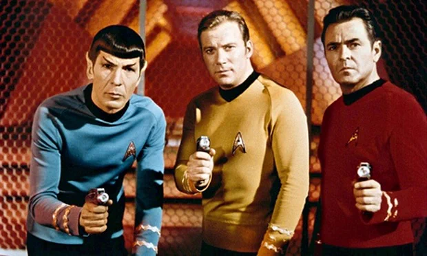 Three men, one wearing blue, one wearing gold, one wearing red, pointing their phasers at the camera. The three men are Spock, Kirk and Scotty.