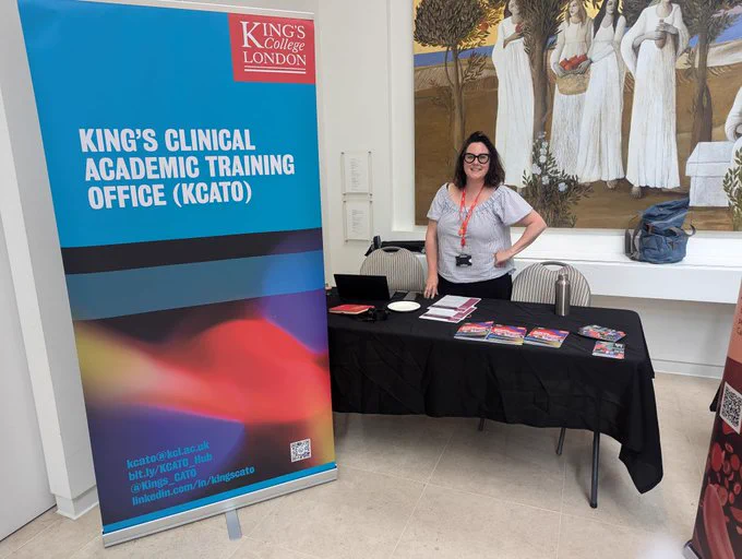 Roll up banner with the words King's Clinical Academic Training Office (KCATO) next to an exhibition table with brochures and a member of the KCATO team smiling.