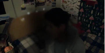 Image shows still from a Macular Society film. The image is of a teenage boy sat on his bed, the image is distorted to represent what it looks like to have macular disease. 