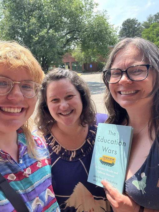 Linds, a blonde non-binary person with glasses and a colorful shirt, with Zandra and Sarah, two women with brown hair wearing dark blue dresses, smile at the camera while Sarah is holding a copy of a blue book titled “The Education Wars” with a yellow school bus on the front cover