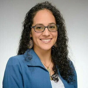 A professional headshot of an individual wearing glasses and a blue blazer, with curly hair and a friendly smile.