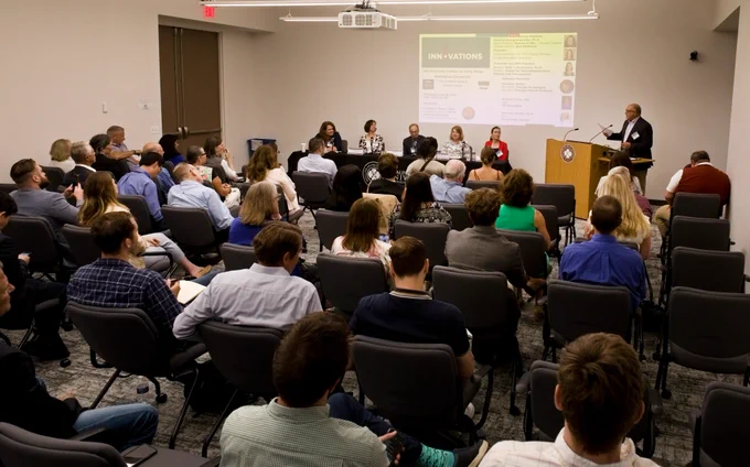 A presentation room filled with attendees and a table of panelists.
