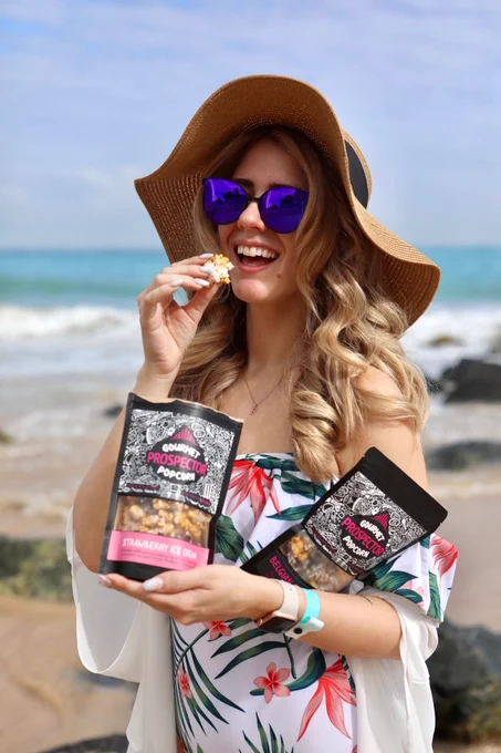 Ren eats a bag of Strawberry Ice Cream gourmet popcorn on a beach while holding a bag of Belgian Chocolate Toffee. She's wearing sunglasses and a floppy hat. Waves can be seen behind her.