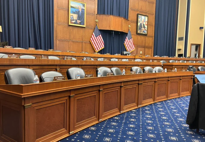 Interior view of a United States congressional hearing room with rows of empty seats and desks, each with a nameplate.