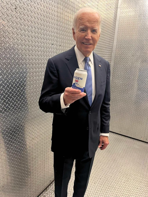 President Joe Biden, dressed in a dark suit and tie, smiles while holding a can. 