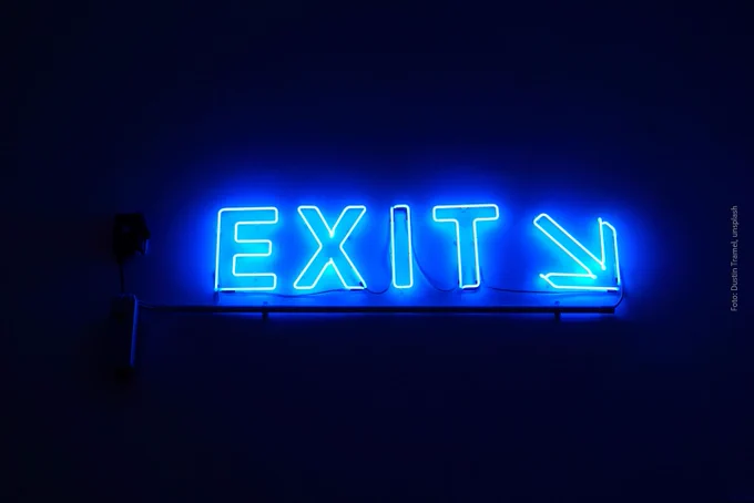 Photo: Dustin Tramel, Unsplash. Photo of an emergency exit sign. Black background. The word "EXIT" in blue illuminated letters in the foreground.