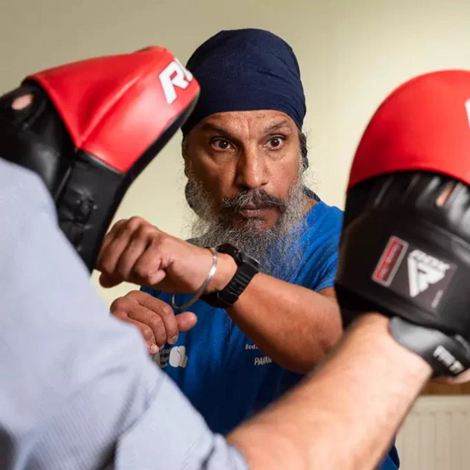 An individual in a blue shirt and turban closely observing two people practicing boxing, each wearing red protective headgear.