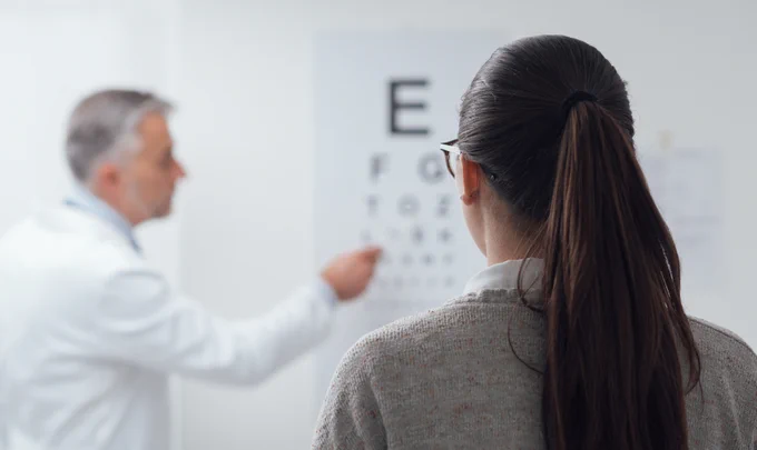 Woman having eye test reading letters from a chart. A man is pointing to the letters she is reading in the background.