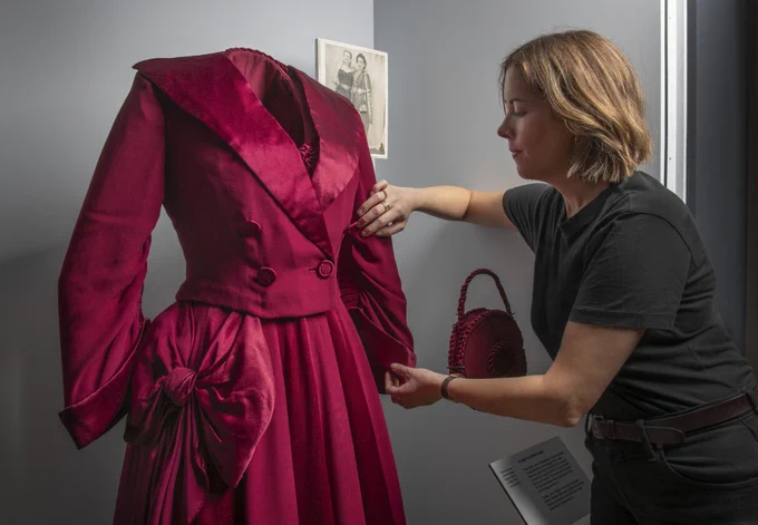 A curator carefully examines a historical deep red dress on a mannequin in a museum exhibit, highlighting fashion history; a black-and-white photograph is visible in the background, adding context to the display.