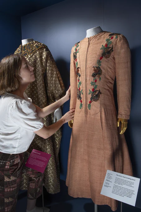 A person examines a vintage embroidered dress on display in a museum exhibit, alongside other historic garments.