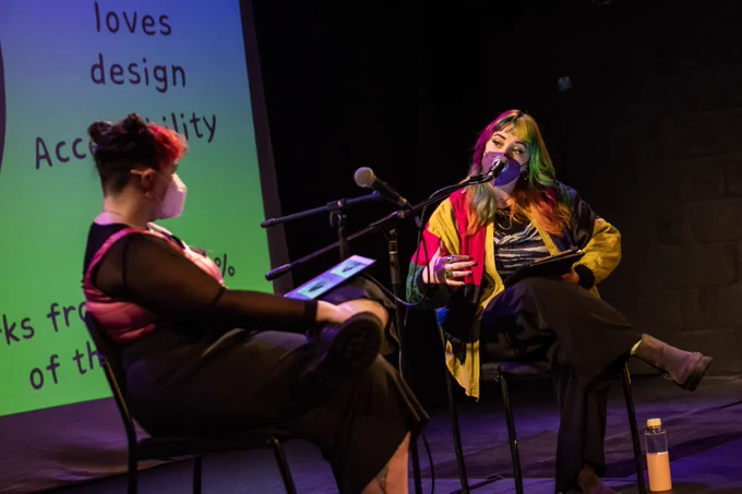 A photo of Tara and Aine sitting towards each other expressively talking as they do a casual presentation and discussion to a full room of people at Project Arts Centre. A large pink and green projection with text behind them. They both wear masks and vibrant clothes in their signature colours green and pink.