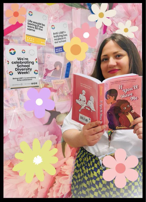 A woman holding a copy of ‘If you’ll have me” a graphic novel by Eunnie, showing the cover and smiling. Background is a school diversity week poster display. Photo has extra flower sticker images all over it.