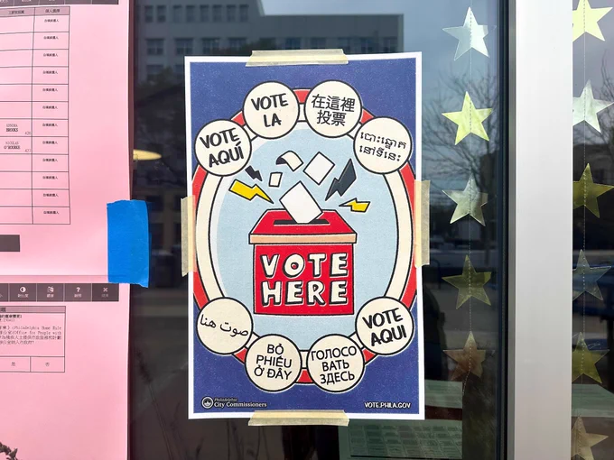 A sign is taped to a glass door, displaying "Vote Here" in multiple languages inside speech bubbles. The background includes reflections and star-shaped decals.