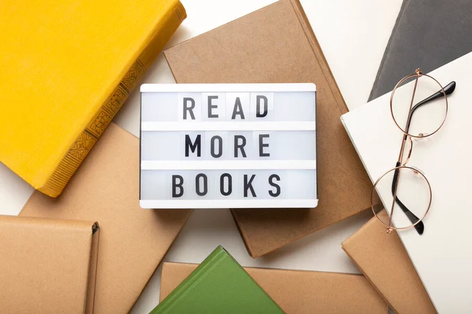 Lightbox with the text "READ MORE BOOKS" surrounded by several books and a pair of glasses, set on a flat surface with various colored hardcover books both opened and closed.