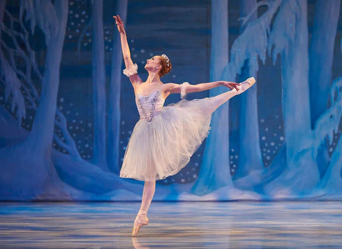A ballet dancer in a white tutu performs on stage, gracefully posing with one leg lifted and pointed, arms elegantly extended. The backdrop depicts a snowy, forest-like scene, enhancing the wintry ambiance of the performance.