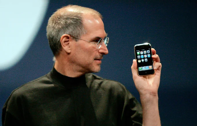 Steve Jobs sosteniendo el primer iPhone. Lo está mirando hacia la izquierda.