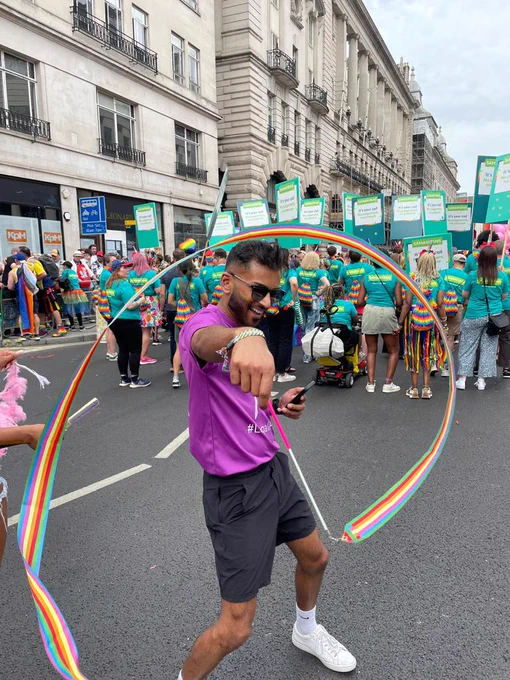 A man in a purple t-shirt spinning a rainbow ribbon. Behind him people are walking in the pride march.