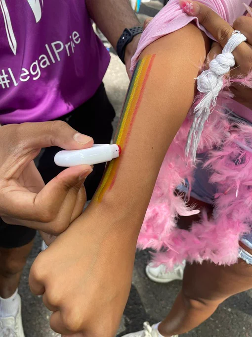 A close up of a person in a purple t-shirt that says #LegalPride putting a rainbow stripe on the arm of another person in pink feathers