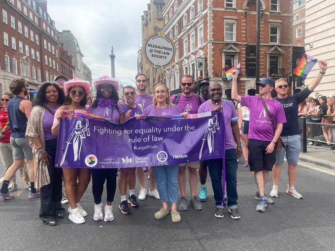 A group of men and women in purple t-shirts holding rainbow flags and holding a large purple banner that reads: “Fighting for equality under the rule of law #LegalPride”