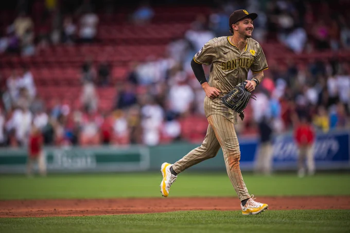 Photo of Jackson Merrill smiling as he runs towards the Padres post-game high five line. He is wearing the Padres sand road pinstripes uniform.