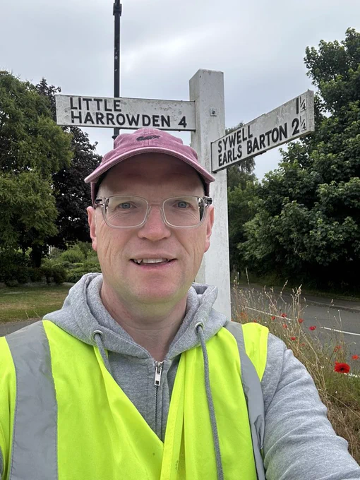 Image shows the author wearing a pink cap, grey hoodie and fluorescent vest stood in front of a directional sign. Pointing left reads Little Harrowden 4, pointing towards the author and over their shoulder reads Sywell 1 Earls Barton 2. At the base of the sign are tall flowers and poppies. The sky is grey.