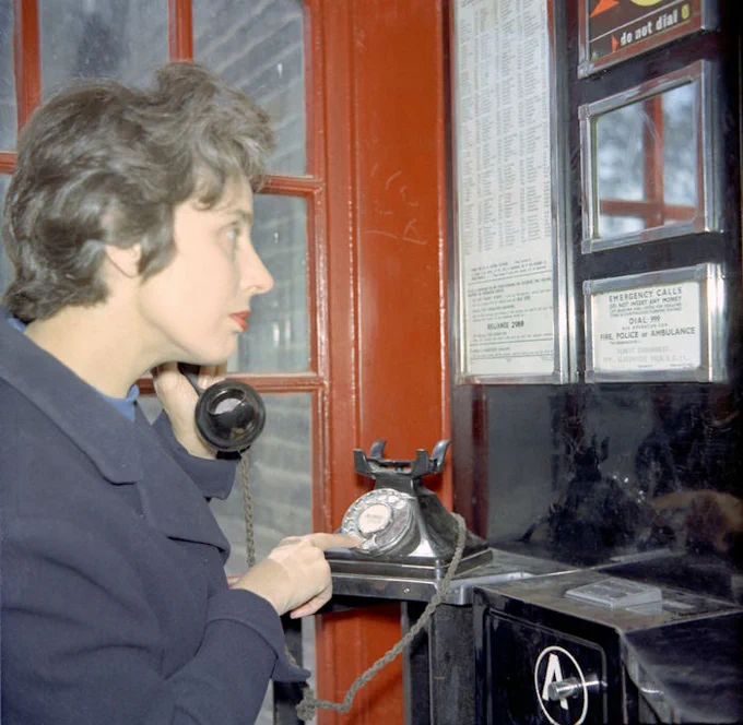 A women is demonstrating the 999 emergency call system using a phone box, taken in the 1950s