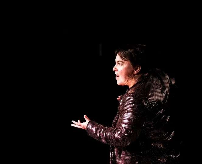 Stephe in the middle of a storytelling performance. She faces to the left, her left hand raised, and her hand open, palm-up. She wears a bronze sequinned jacket, which is reflecting the stage lights onto her skin.