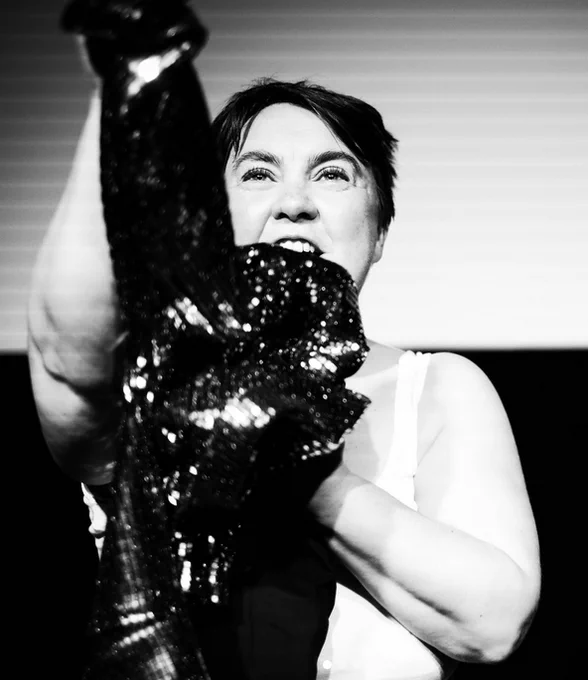Stephe in the middle of a storytelling performance. She's facing front, and speaking vehemently. She wears a white vest, and is gripping a sequinned blazer to her chest.