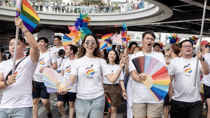 Team members march with flags, fans, and pinwheels during the Bangkok Pride Parade.