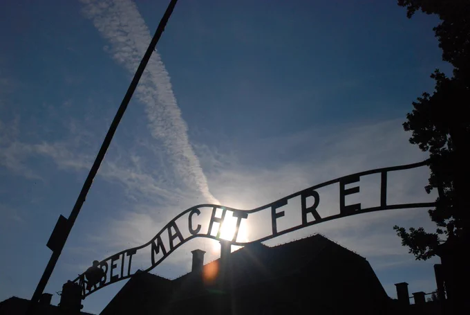 The Arbeit macht frei sign from the Auschwitz I site (contemporary view). The inscription is photographed against the sky. The sun and clouds are visible as well as silhouettes of buildings and trees.