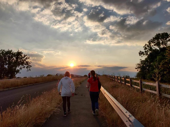 Two people walking away from the camera towards a sunset. They are on a path bordered by long grass, which crosses over a motorway bridge.