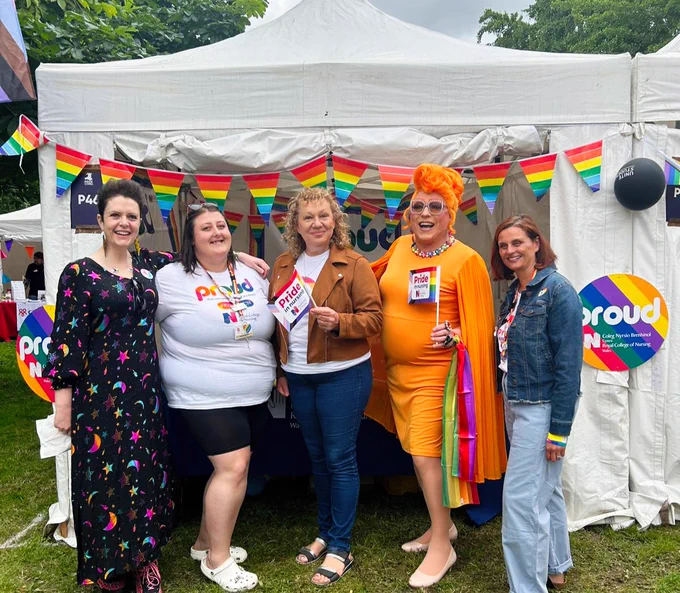 Five people stand together in front of a "Proud" booth adorned with rainbow flags at a Pride celebration in Cardiff, Wales.