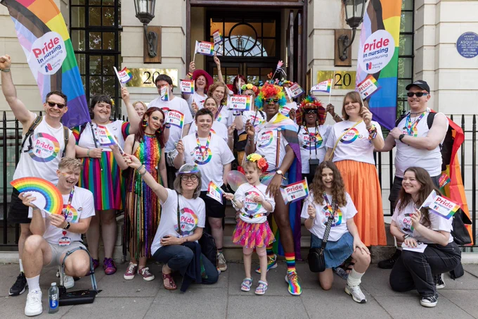 A group of RCN members and staff celebrating at a Pride event in front of the Royal College of Nursing HQ in London, with banners displaying "Pride" and rainbow flags. Individuals are wearing colourful attire and accessories.