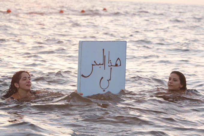 Photo of two people in the sea, which occupies the whole frame. We see only their heads, both of them with long brown hair. They are holding above the water a placard with the phrase ‘This Sea is Mine’ written in Arabic.