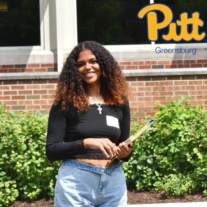 Student standing outside smiling, holding folder. Pitt-Greensburg script logo in top right corner of image.