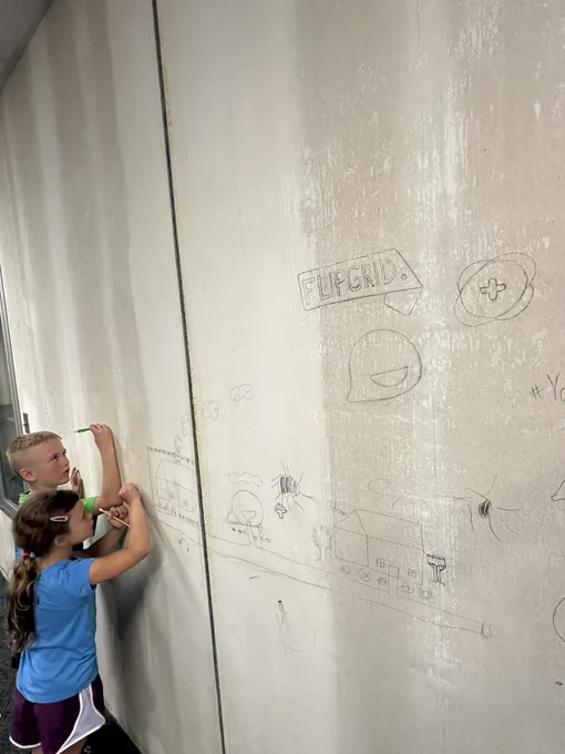Flipgrid tag logo, Plus symbol logo, and Smubble logo drawn on a wall. Two kids drawing on the wall drawing a neighborhood with buildings and even a Smubble building.