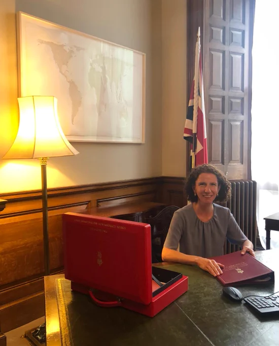 Anneliese Dodds sits at her new desk as Minister for International Development, holding a red ministerial folder and with a red box in front of her. In the background a map of the world is visible.