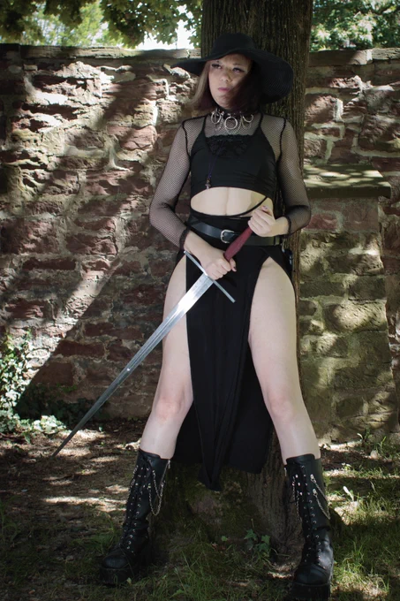 A photoshoot photo of me, Flora, wearing a black skirt that shows my legs through 2 splits, a black crop top with mesh sleeves and a big black hat. I hold a long sword upside down and lean against a tree in a firm, broad pose, looking into the distance